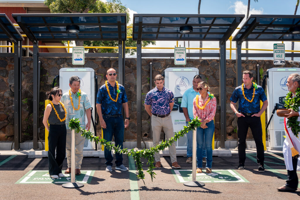 Project partners, community leaders, and attendees gather behind a ceremonial ti leaf rope in anticipation of the kahu's blessing of the new EV charging station at Kapalua Airport, honoring Hawaiian cultural traditions and the project's opening.