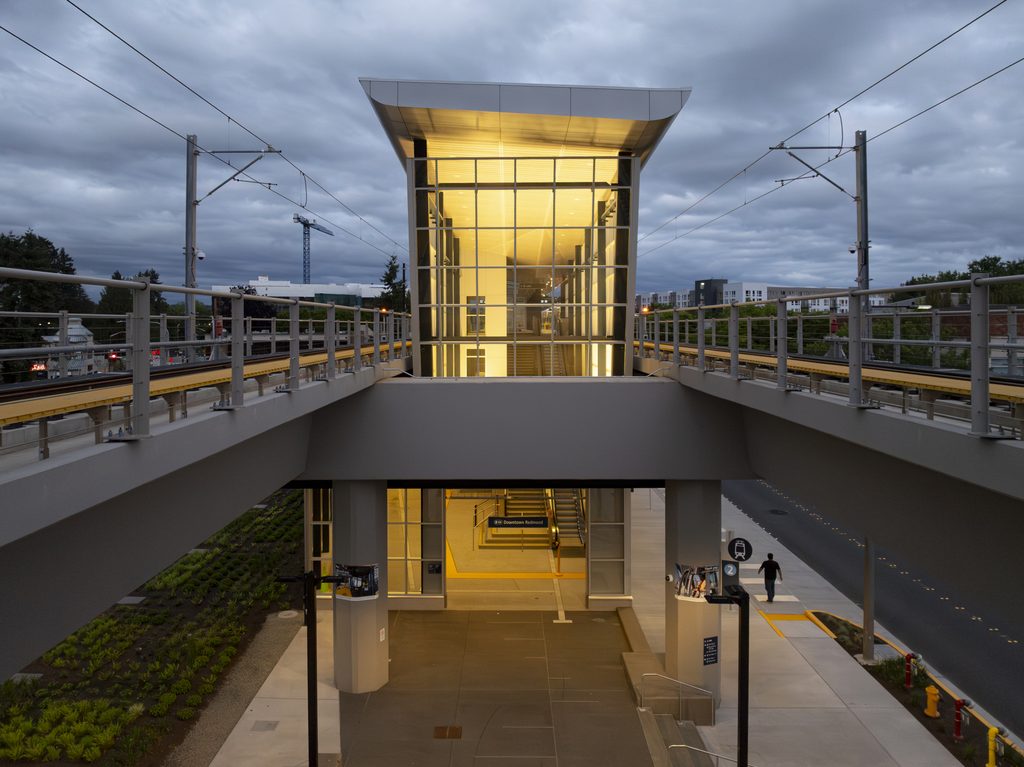 Elevated Downtown Redmond Link Extension station with a brightly lit glass entrance, light rail tracks and overhead power lines beneath a cloudy evening sky.