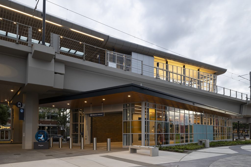 Downtown Redmond Link Extension station entrance beneath elevated light rail tracks, featuring a glass-enclosed lobby, covered walkway and illuminated platform above.